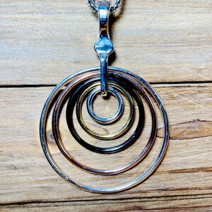 May include: A silver chain necklace with a pendant featuring five concentric circles in silver, rose gold, black, gold, and silver.
