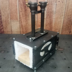 May include: A black metal forge with a white refractory lining and two gas inlets. The forge is sitting on a metal surface.