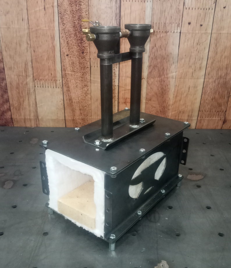 May include: A black metal forge with a white refractory lining and two gas inlets. The forge is sitting on a metal surface.