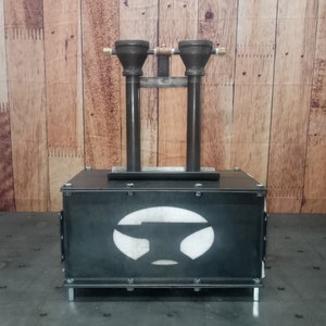 May include: A black metal forge with two vertical pipes and a white anvil logo. The forge is sitting on a black surface.