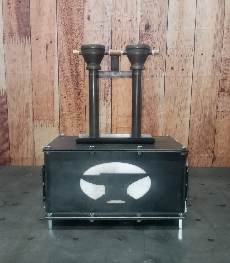 May include: A black metal forge with two vertical pipes and a white anvil logo. The forge is sitting on a black surface.