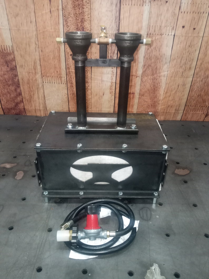 May include: A black metal forge with a cutout of a hammer in the front. The forge has two gas burners with a black hose and regulator.