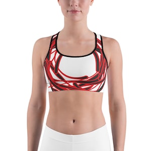 May include: A white sports bra with a black band and red abstract design. The design features a circle with red lines and black lines.