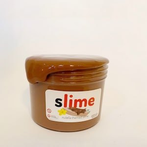 Puede incluir: Una slime marr&oacute;n en un tarro transparente con una etiqueta que dice "slime" en letras rojas y "nutella themed slime" en letras negras.