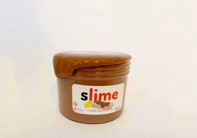 Puede incluir: Una slime marr&oacute;n en un tarro transparente con una etiqueta que dice "slime" en letras rojas y "nutella themed slime" en letras negras.