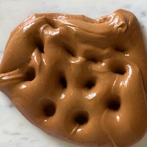 Puede incluir: Primer plano de slime marr&oacute;n con un acabado brillante y varios huecos de aire. La slime est&aacute; sobre una superficie de m&aacute;rmol blanco.