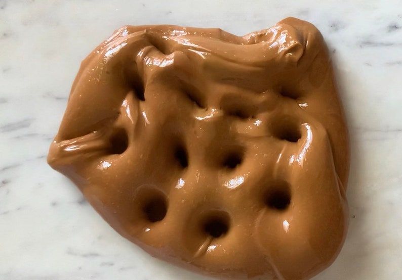 Puede incluir: Primer plano de slime marr&oacute;n con un acabado brillante y varios huecos de aire. La slime est&aacute; sobre una superficie de m&aacute;rmol blanco.