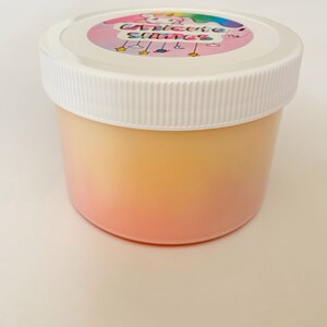 May include: A clear plastic jar with a white lid. The jar contains a pink and yellow ombre slime. The lid has a sticker with a cartoon unicorn and the text "Unicorns Slime".