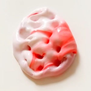 May include: A close-up of a blob of pink and white slime. The slime is a mix of light pink and white, with streaks of bright red. The texture appears smooth and glossy, with small holes throughout. The background is plain white.