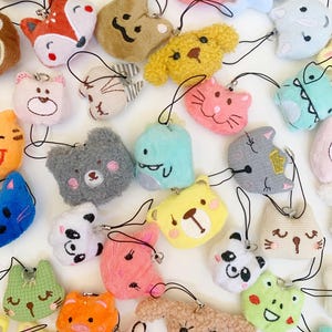 Puede incluir: Una colección de llaveros de animales de peluche coloridos con caras bordadas. Los llaveros presentan una variedad de animales, incluidos gatos, perros, osos, pandas, zorros y más.