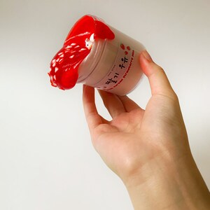 May include: A clear plastic pot filled with pale pink slime, overflowing with bright red slime. The pot has text in Korean and English, "with Korean Strawberry Milk". A small white figure is partially visible within the red slime.