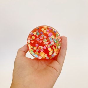 Può includere: Un sottobicchiere rotondo, arancione e trasparente in resina con una varietà di ciondoli di frutta e caramelle colorati incorporati al suo interno.