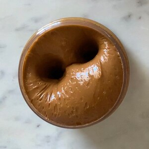 Puede incluir: Primer plano de una mantequilla de cacahuete marr&oacute;n en un tarro de vidrio. La mantequilla de cacahuete es suave y cremosa, con un remolino en el centro.