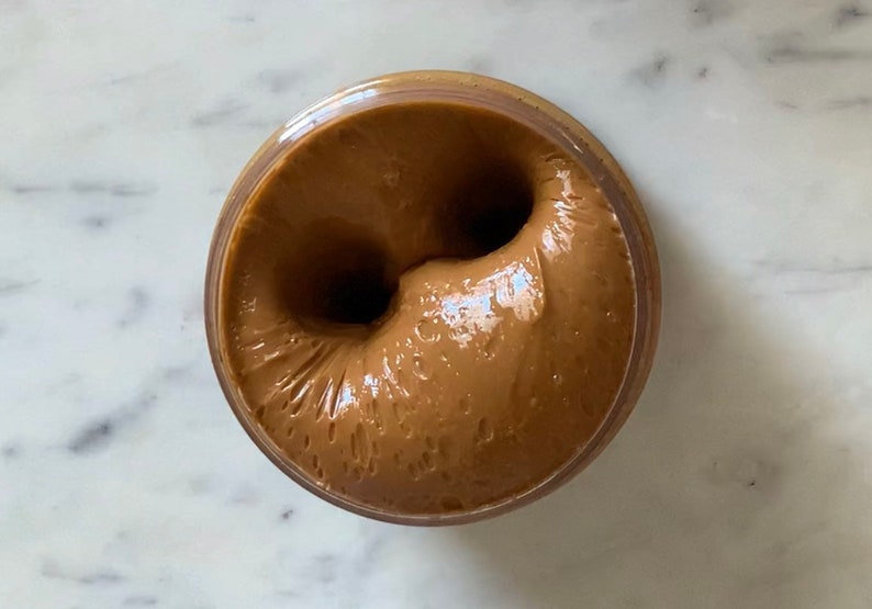 Puede incluir: Primer plano de una mantequilla de cacahuete marr&oacute;n en un tarro de vidrio. La mantequilla de cacahuete es suave y cremosa, con un remolino en el centro.