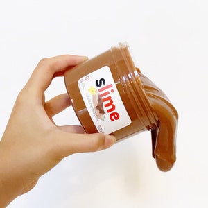 Puede incluir: Una slime marr&oacute;n en un tarro transparente con una etiqueta blanca que dice "slime" en letras rojas y "Nutella themed slime" en letras negras.