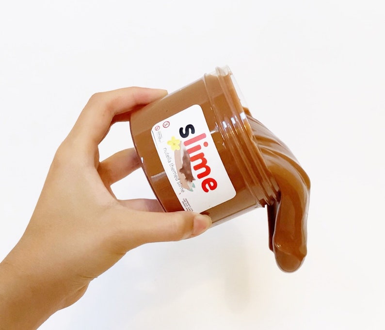 Puede incluir: Una slime marr&oacute;n en un tarro transparente con una etiqueta blanca que dice "slime" en letras rojas y "Nutella themed slime" en letras negras.