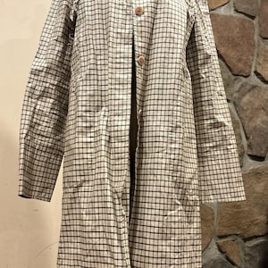 May include: A long-sleeved, knee-length coat featuring a beige and dark blue grid pattern. The coat has a pointed collar, a front button closure with four visible round brown buttons, and long sleeves. It is displayed on a mannequin torso.