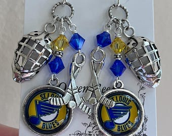 May include: Silver-toned hockey-themed earrings. Each earring features a hockey mask charm, a St. Louis Blues logo charm, and blue and yellow beads. The logo charm has a blue note and hockey stick design.