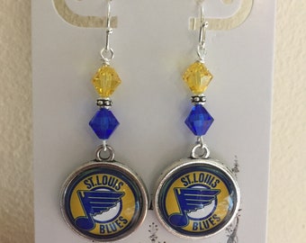 May include: A pair of silver dangle earrings featuring a St. Louis Blues hockey team logo charm. The charms are round and have a blue, yellow, and white design. The earrings are accented with blue and yellow crystal beads.
