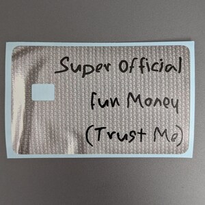 May include: A rectangular sticker with a metallic silver, textured surface. The text "Super Official Fun Money (Trust Me)" is written in black marker across the center. A small, plain white rectangular area is visible on the left side.