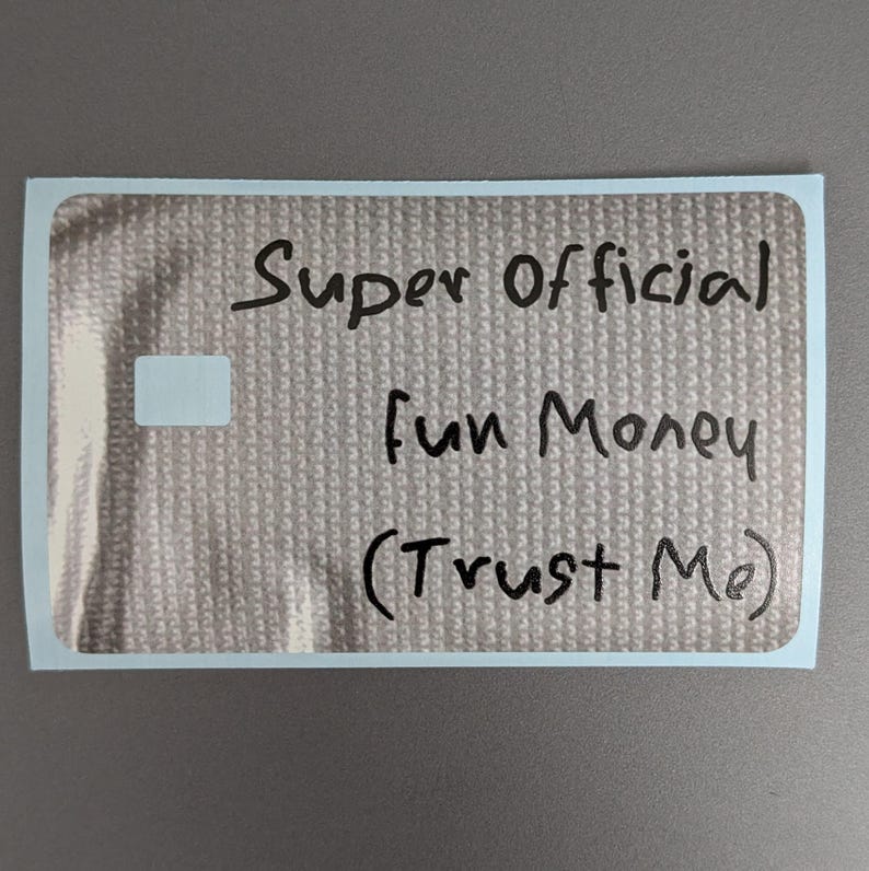 May include: A rectangular sticker with a metallic silver, textured surface. The text "Super Official Fun Money (Trust Me)" is written in black marker across the center. A small, plain white rectangular area is visible on the left side.