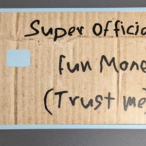 May include: A rectangular card with a light blue border and a cardboard texture. The card has handwritten text in black ink that reads "Super Official fun Money (Trust me)" and a small light blue square.