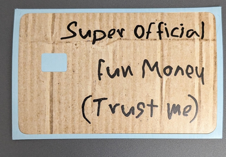 May include: A rectangular card with a light blue border and a cardboard texture. The card has handwritten text in black ink that reads "Super Official fun Money (Trust me)" and a small light blue square.