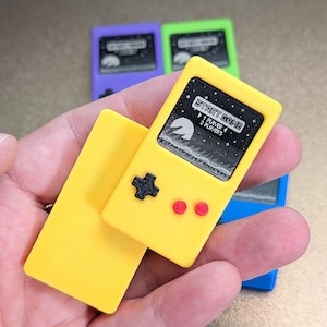 May include: A yellow miniature handheld game console with a black directional pad and red buttons. The screen displays a pixelated graphic with the text "START GAME" and "1 PLAYER 2 PLAYERS". Other consoles in purple, green, and blue are visible in the background.