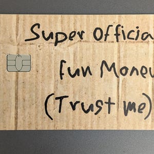 May include: A rectangular card made of corrugated cardboard with rounded corners. The card has the words "Super Official fun Money (Trust me)" written in black marker. A silver SIM card is on the left side.