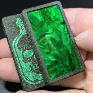 May include: Two rectangular, dark green objects. One features a green skull and bottle design, the other has a swirling, marbled green interior. The objects are held in a hand, showcasing their contrasting textures and colors.