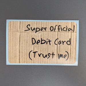Puede incluir: Una pegatina rectangular con un borde azul claro que presenta un fondo de textura de cart&oacute;n marr&oacute;n. El texto "Super Official Debit Card (Trust me)" est&aacute; escrito con rotulador negro en el centro de la superficie de cart&oacute;n con un estilo manuscrito.