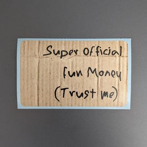 Puede incluir: Una pegatina rectangular con un borde azul claro que presenta un fondo con textura de cart&oacute;n marr&oacute;n. El texto Super Official Fun Money (Trust me) est&aacute; escrito con marcador negro en el centro de la pegatina en un estilo manuscrito informal.
