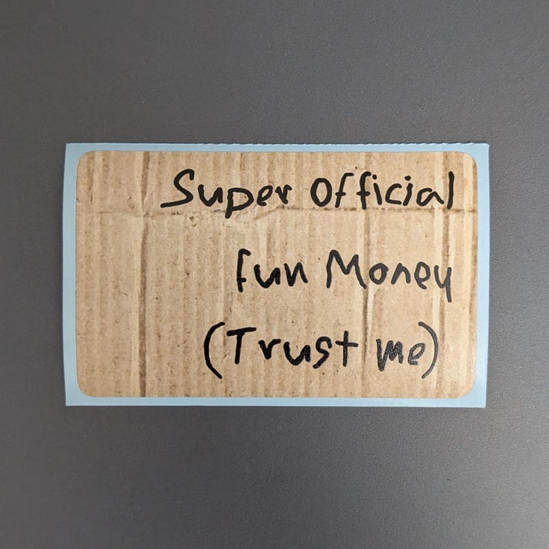 Puede incluir: Una pegatina rectangular con un borde azul claro que presenta un fondo con textura de cart&oacute;n marr&oacute;n. El texto Super Official Fun Money (Trust me) est&aacute; escrito con marcador negro en el centro de la pegatina en un estilo manuscrito informal.