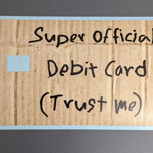 May include: A novelty debit card made from cardboard, with the words "Super Official Debit Card (Trust me)" written in black marker. The card has a light blue border and a small, light blue square cutout.