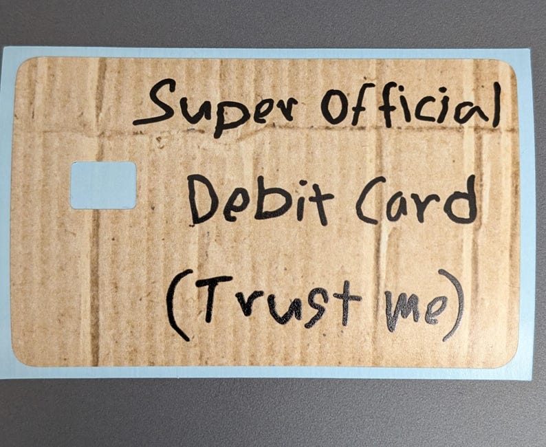 May include: A novelty debit card made from cardboard, with the words "Super Official Debit Card (Trust me)" written in black marker. The card has a light blue border and a small, light blue square cutout.