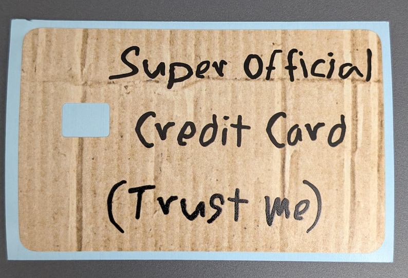May include: A rectangular card with a light blue border and a cardboard-like texture. The text "Super Official Credit Card (Trust me)" is handwritten in black. A small, light blue rectangle is in the upper left corner.