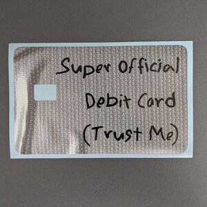 May include: A rectangular sticker with a metallic silver textured surface. The text "Super Official Debit Card (Trust Me)" is written in black marker across the center. A small, solid white rectangular shape is positioned on the left side.