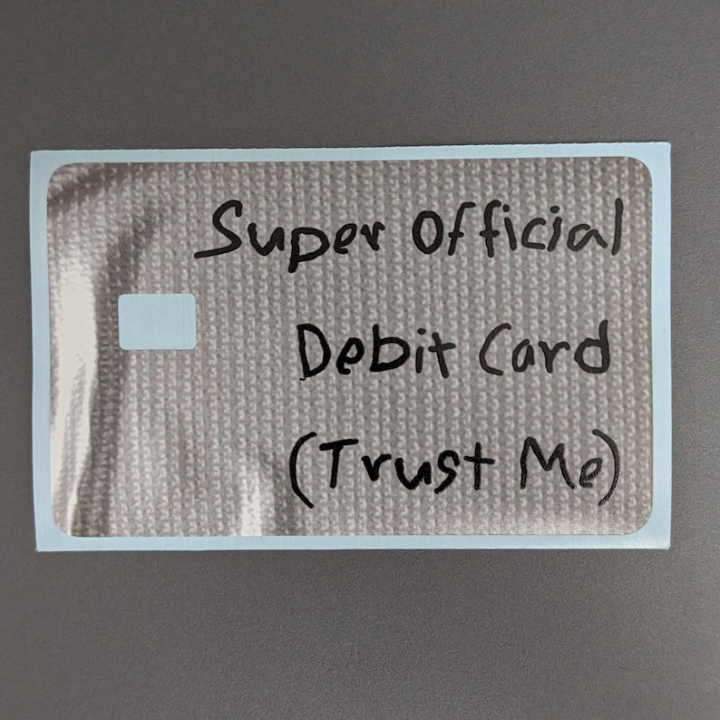 May include: A rectangular sticker with a metallic silver textured surface. The text "Super Official Debit Card (Trust Me)" is written in black marker across the center. A small, solid white rectangular shape is positioned on the left side.