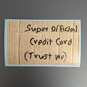 Puede incluir: Una pegatina rectangular con un borde azul claro que presenta un fondo de textura de cart&oacute;n ondulado marr&oacute;n. El texto "Super Official Credit Card (Trust me)" est&aacute; escrito con rotulador negro en el centro de la superficie de cart&oacute;n.