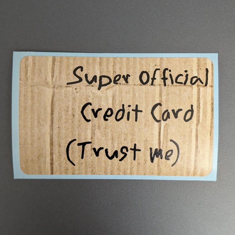 Puede incluir: Una pegatina rectangular con un borde azul claro que presenta un fondo de textura de cart&oacute;n ondulado marr&oacute;n. El texto "Super Official Credit Card (Trust me)" est&aacute; escrito con rotulador negro en el centro de la superficie de cart&oacute;n.