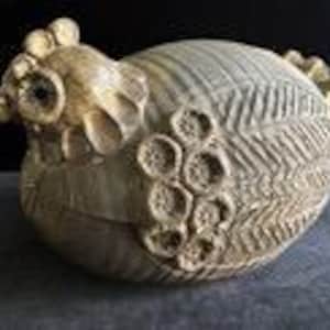May include: A beige ceramic chicken figurine featuring a textured surface with a herringbone pattern on the body and a series of raised, circular floral-like impressions along the wing area, with a defined comb and beak.