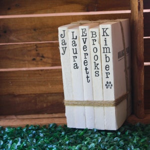 May include: A set of six white books with black lettering, each book has a different name on the spine: Jay, Laura, Everett, Brooks, Kimber, and Miller. The books are stacked and tied together with twine.