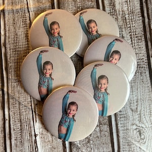 May include: Six round white buttons with a glossy finish. Each button features a photo of a young person wearing a blue and white patterned outfit, with their arm raised in a dance pose.