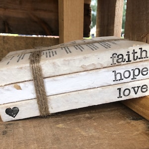 May include: Three white books stacked on top of each other. The books are tied together with twine. The top book has the word "faith" printed on it. The middle book has the word "hope" printed on it. The bottom book has the word "love" printed on it. A small black heart is printed on the bottom book.