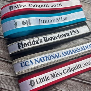 May include: Five stacked pageant sashes featuring various colors including red, teal, black, and white. The sashes display text such as Miss Colquitt 2026, Junior Miss 2026 NC Poultry Jubilee, Florida's Hometown USA, UCA NATIONAL AMBASSADOR, and Little Miss Colquitt 2026.