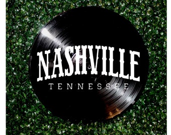 May include: A black vinyl record featuring the word "NASHVILLE" in white, with "TENNESSEE" underneath. The record is displayed on a backdrop of dense, green foliage.