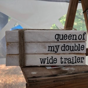 May include: Three white wooden blocks stacked on top of each other, tied together with twine. The blocks have black lettering that reads "queen of my double wide trailer".
