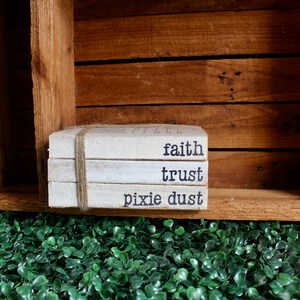 May include: A stack of three white books with the words "faith", "trust", and "pixie dust" printed on them. The books are tied together with twine and are sitting on a wooden shelf.