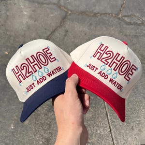May include: Two baseball caps with off-white crowns and embroidered text reading H2HOE JUST ADD WATER in red with light blue water drop accents. One cap features a navy blue brim, while the other cap features a bright red brim.