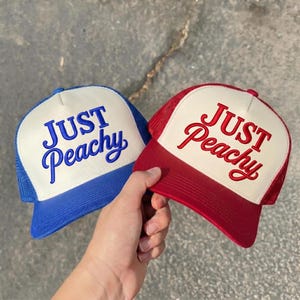 May include: Two trucker hats with white front panels and mesh backs. One hat features a blue brim and blue embroidered text that reads Just Peachy. The other hat features a red brim and red embroidered text that reads Just Peachy.